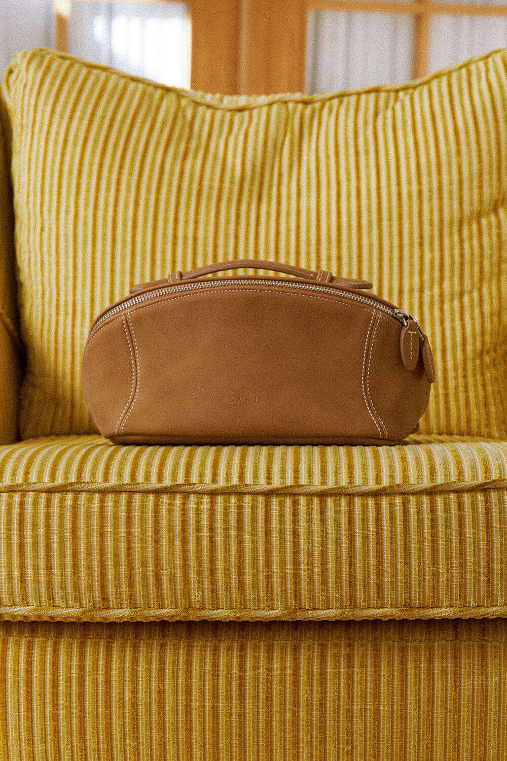 Camel brown makeup bag resting on yellow sofa, retro style travel cosmetic bag.